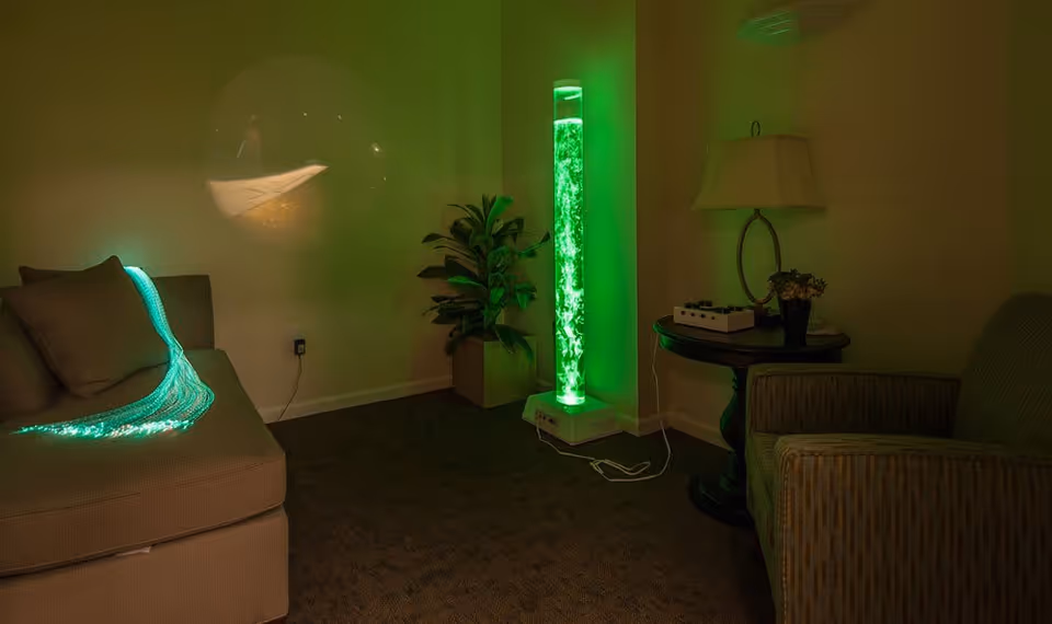A dimly lit room with a beige couch on the left, a green glowing fiber optic lamp draped over the couch, a tall cylindrical bubble lamp emitting green light in the corner, a potted plant next to the lamp, and a round wooden side table with a lamp and small decorative items on the right, along with a cushioned armchair.