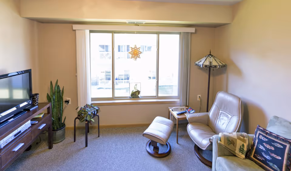 A cozy living room with a large window letting in natural light. The room features a leather recliner with matching ottoman, a small side table with magazines, a floor lamp with a stained glass shade, a green armchair with decorative pillows, a wooden TV stand with a flat-screen TV, and two potted plants on the floor.