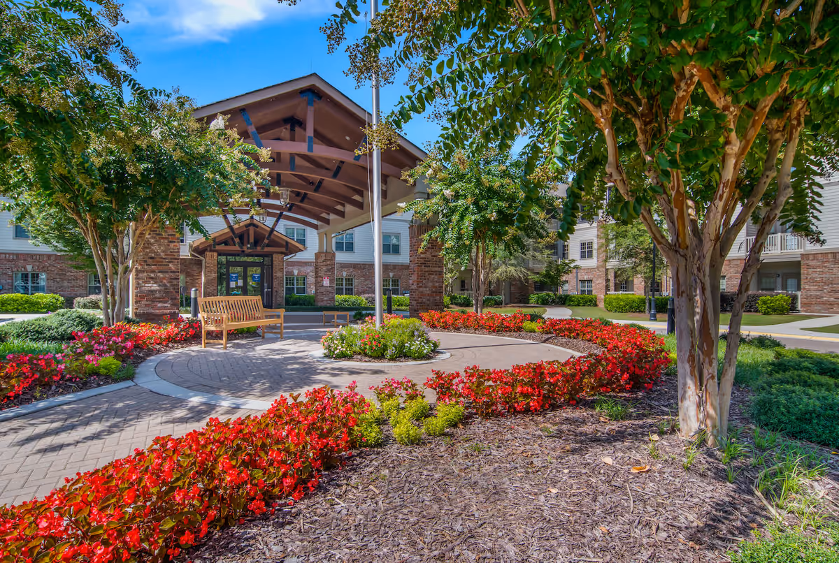 Covered entrance to a senior living facility with a wooden bench, circular brick walkway and landscaped beds of red flowers and trees.