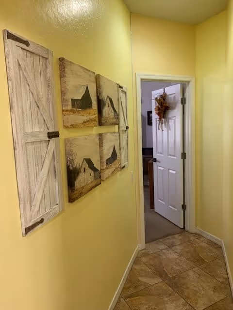 A narrow hallway with yellow walls and tiled floor leading to an open white door decorated with a fall-themed wreath. On the left wall, there are six rustic-style wall decorations, including two barn door panels and four pictures of barns and rural scenes.