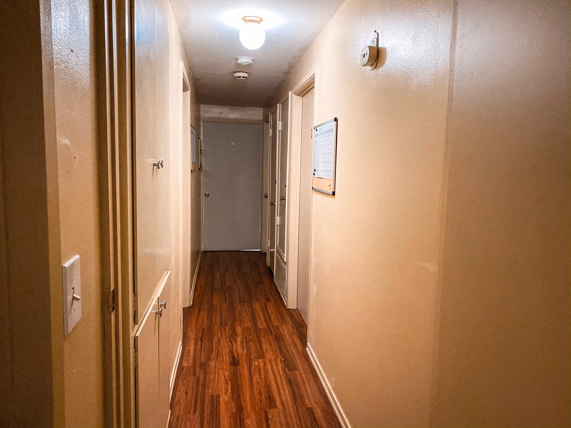 A narrow hallway with wooden flooring and beige walls, featuring several closed doors on both sides and a ceiling light fixture illuminating the space.