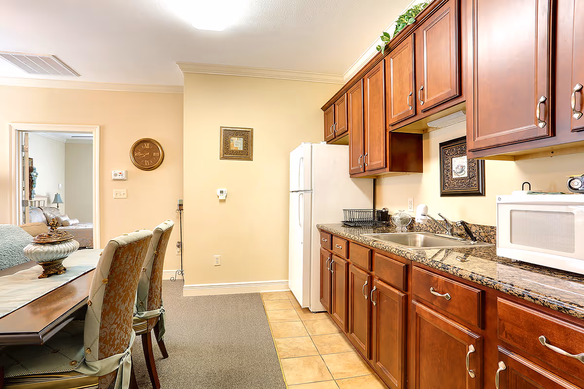 Bright kitchen with wooden cabinets, granite countertops, a sink and microwave, and a dining table with upholstered chairs visible.