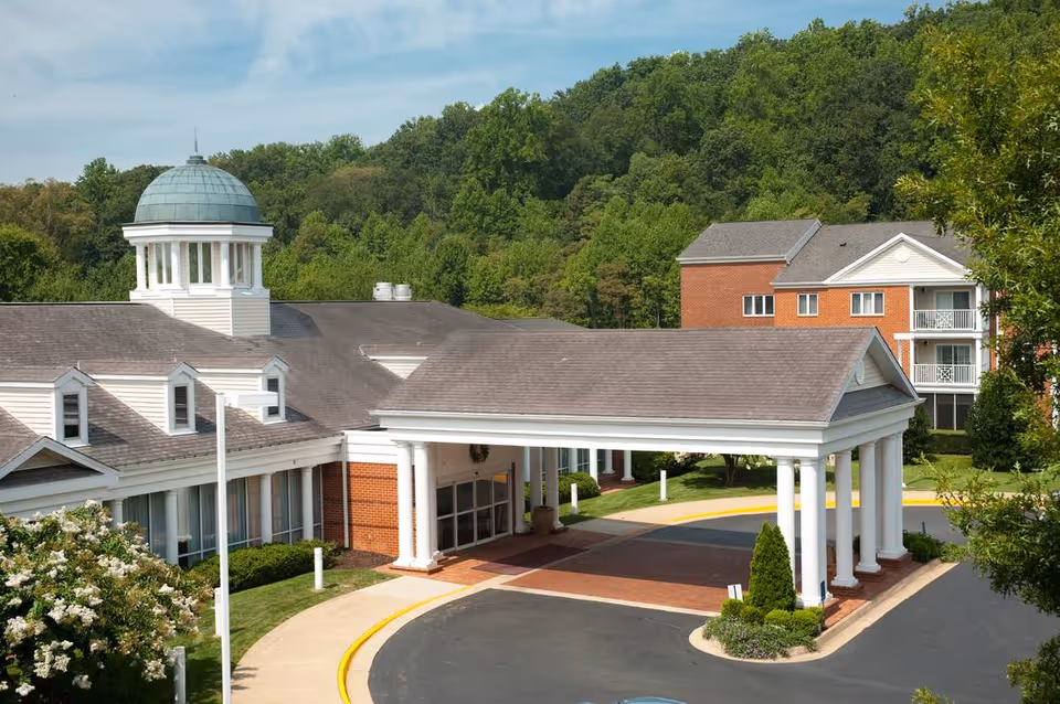 Exterior view of The Colonnades senior living facility showing a covered entrance with white columns, a building with a dome, and surrounding greenery and trees.
