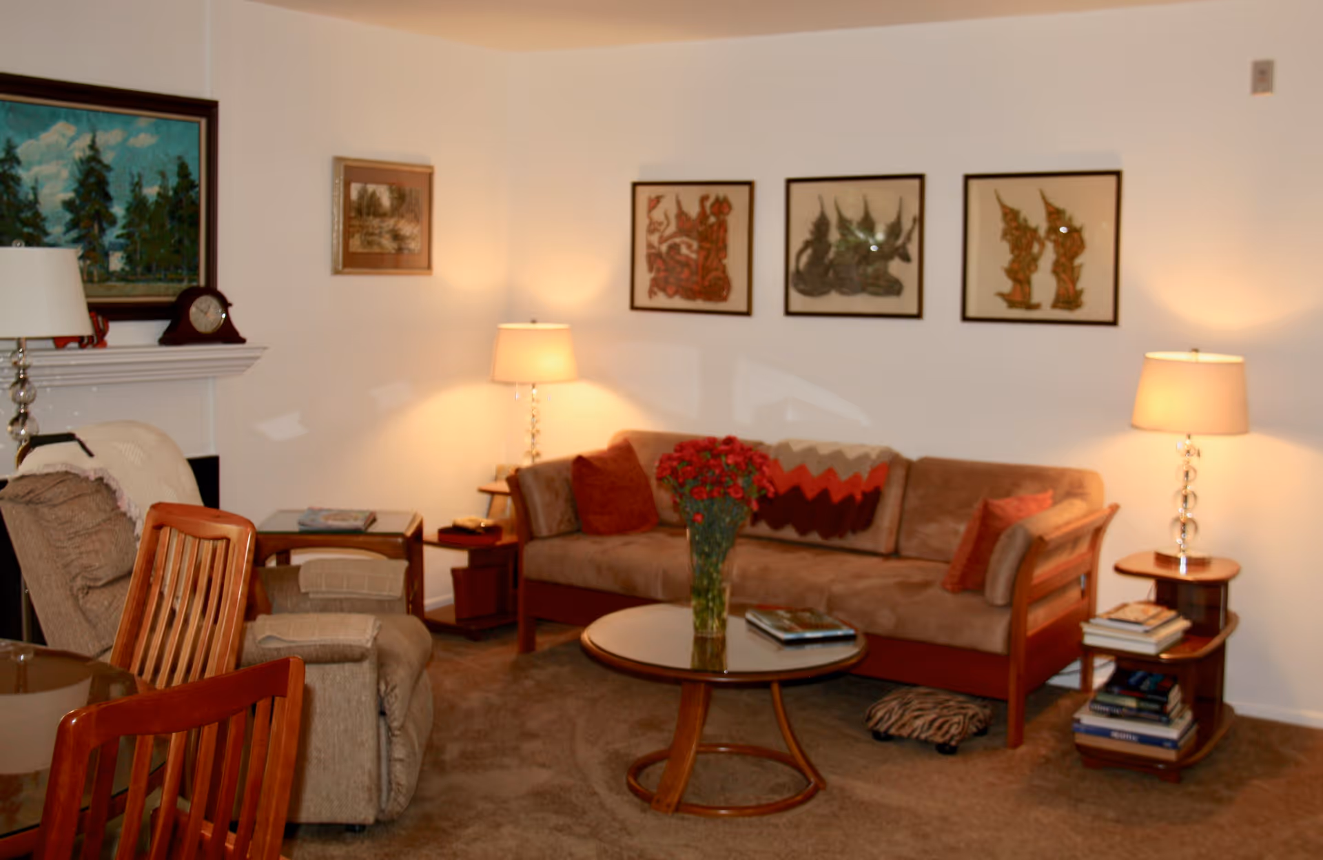 A cozy living room with a brown sofa adorned with cushions and a knitted throw, two side tables each with a lamp, a round glass coffee table with a vase of red flowers, a recliner chair, wooden dining chairs, and framed artwork on the walls. There is also a fireplace with a clock and a painting above it.