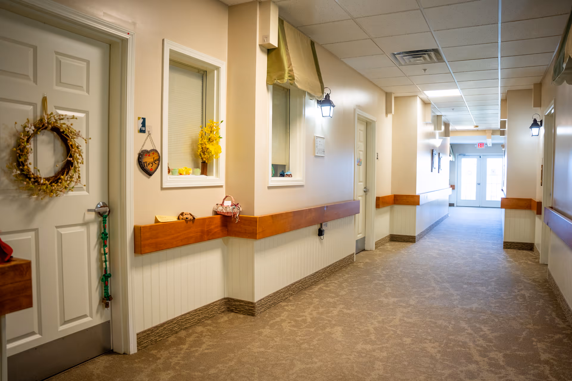 A well-lit hallway in a senior living facility with beige walls, carpeted floor, and wooden handrails. Doors line the hallway, one decorated with a wreath and a hanging sign. Small decorative items and flowers are placed on ledges along the walls. The hallway leads to a set of glass double doors at the far end.