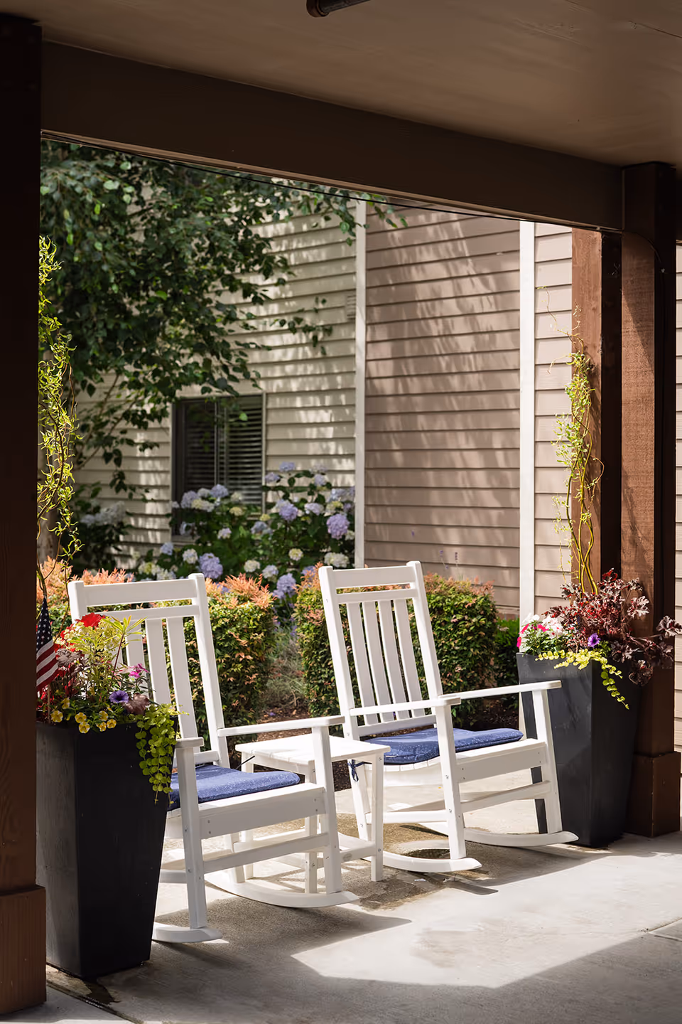 Two white rocking chairs with blue cushions sit on a covered porch between planters in front of a residential building.