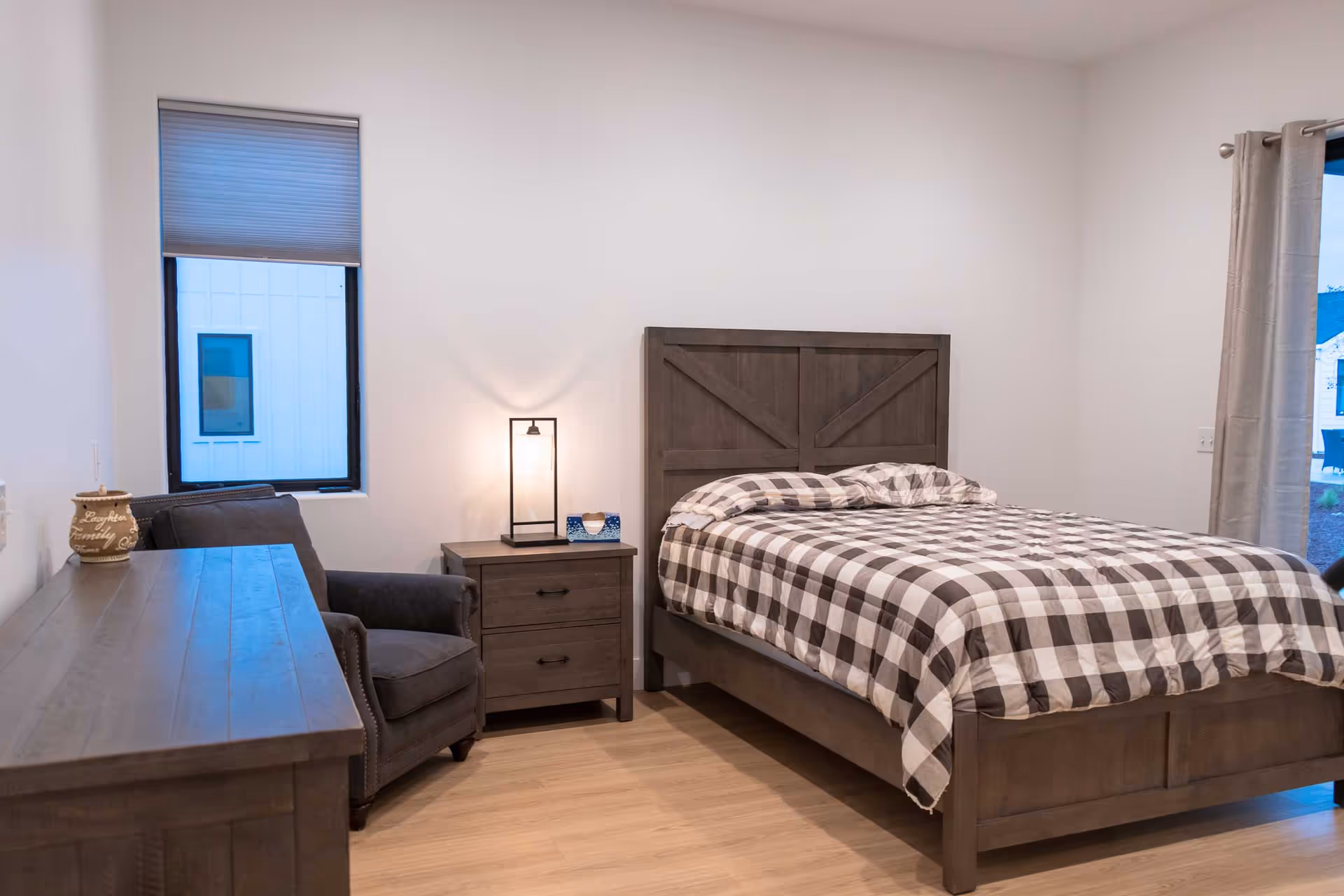 A bedroom with a wooden bed frame and a checkered bedspread. Next to the bed is a wooden nightstand with a modern lamp and a tissue box. There is a gray upholstered armchair beside a wooden dresser. The room has light-colored wooden flooring, white walls, a window with a closed blind, and a sliding glass door with curtains partially open showing an outdoor view.