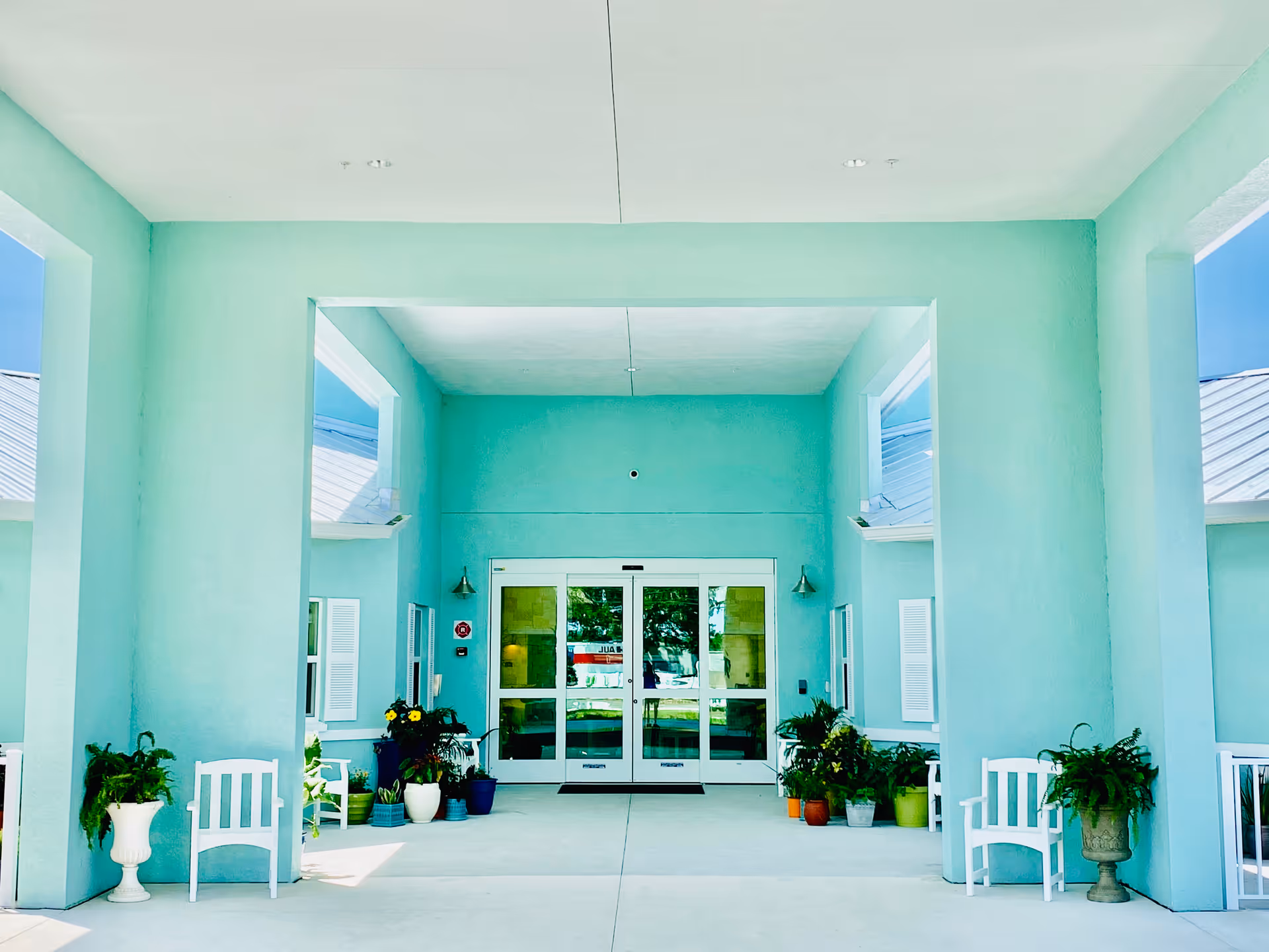 Covered aqua-colored entrance to a building with glass double doors, white benches, and potted plants.