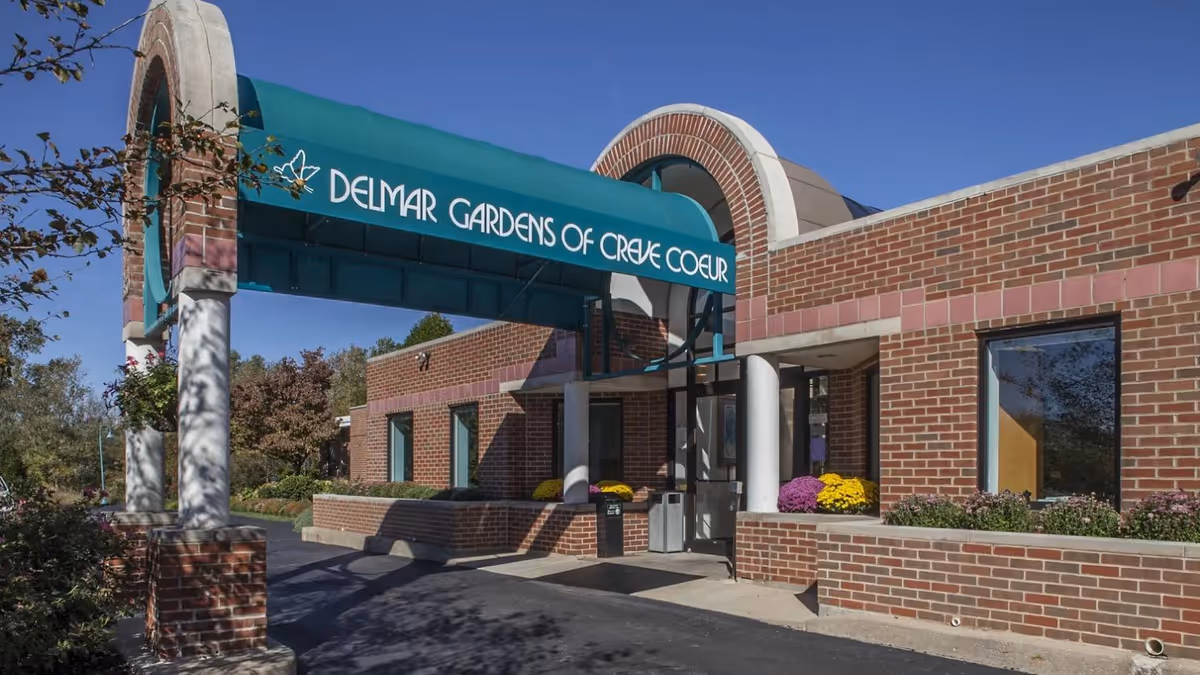 Exterior view of Delmar Gardens of Creve Coeur facility entrance with a brick building and a green awning displaying the facility name. There are flowers planted in front of the building and trees in the background under a clear blue sky.