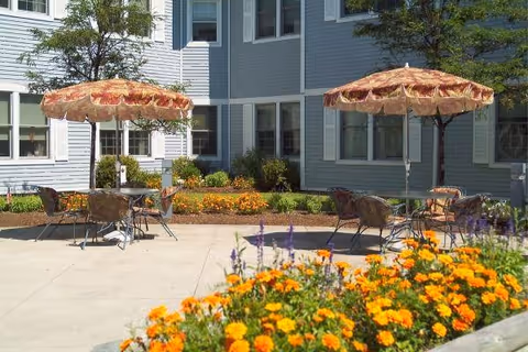 Sunny courtyard with patio tables, fringed umbrellas, and bright flower beds in front of a light-blue nursing facility.