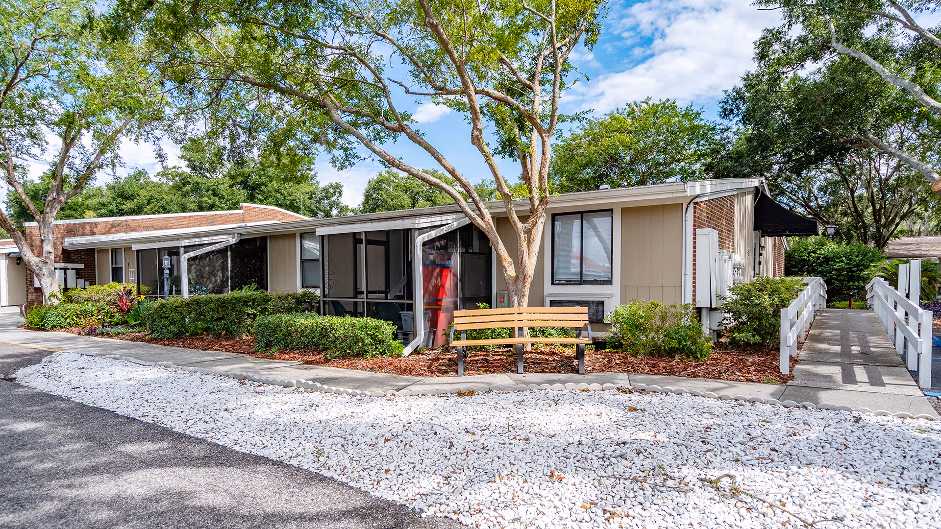 Exterior view of a single-story residential building at Lake Ridge Village with a bench in front, surrounded by trees, bushes, and a white gravel area. There is a ramp leading to the entrance on the right side.