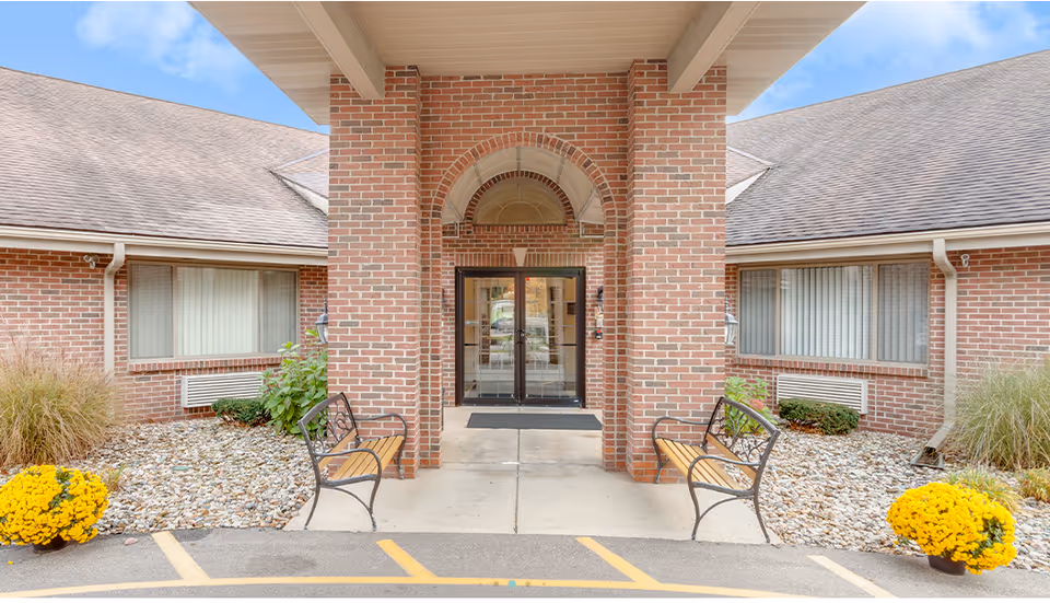 Entrance to Brentwood at Elkhart Assisted Living featuring a brick facade with an arched doorway, double glass doors, two benches on either side of the entrance, decorative plants, and yellow flowers in pots near the walkway.