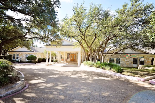 Front entrance of a stone-faced assisted living building with a covered porte-cochère and trees lining the circular driveway.