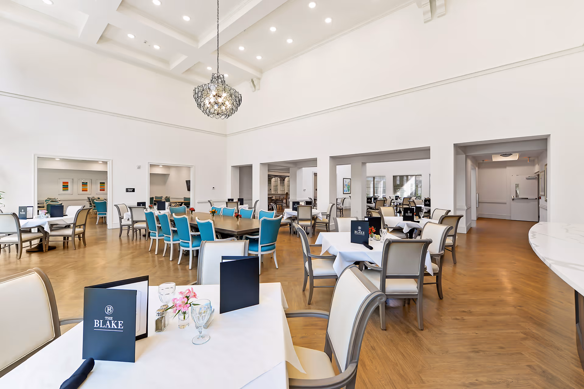 A spacious, well-lit dining room with multiple tables and chairs arranged neatly. The tables are covered with white tablecloths and have menus labeled 'The Blake' along with small flower vases. The room features high ceilings with a decorative light fixture and wooden flooring.