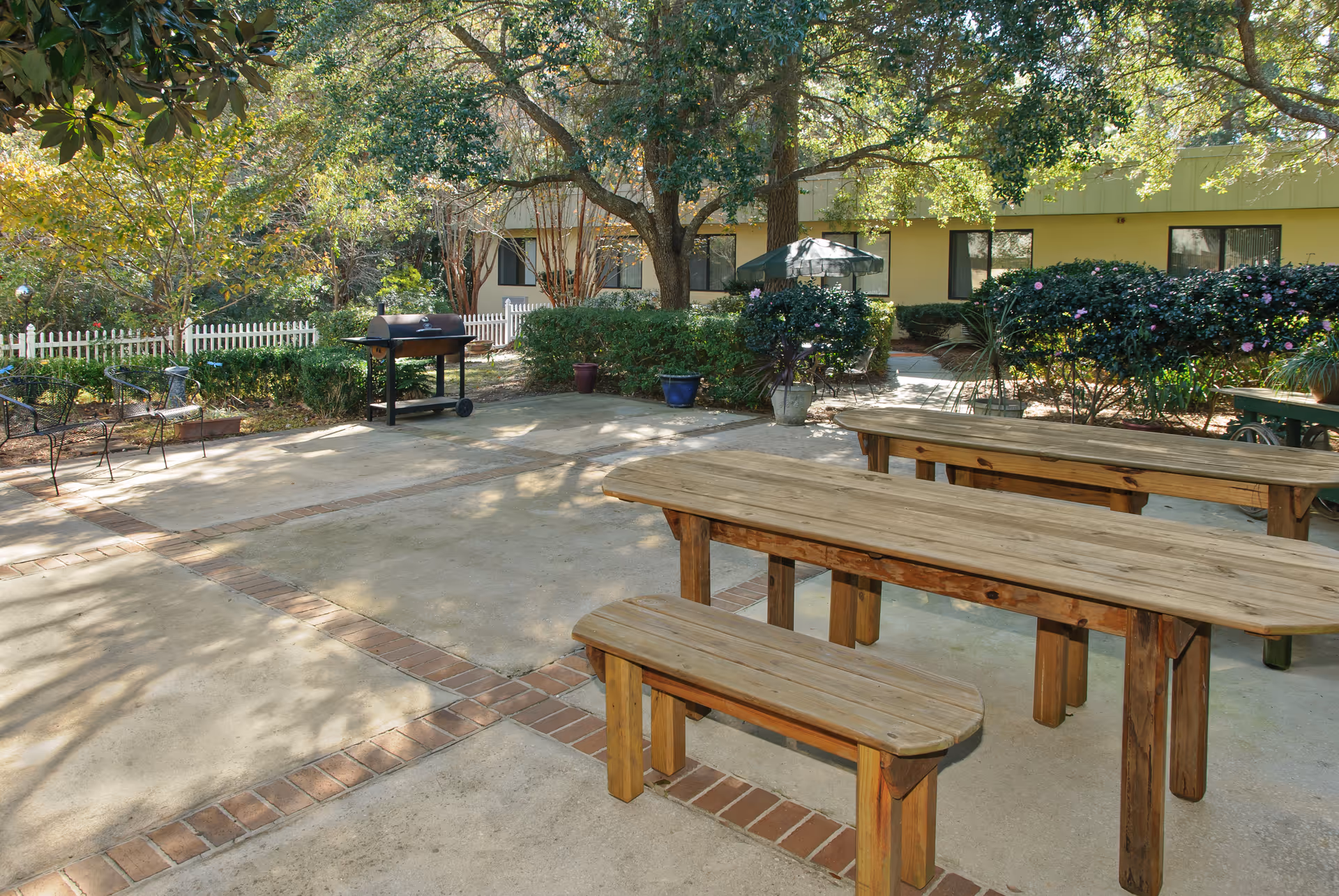 Outdoor patio area with wooden picnic tables and benches, a barbecue grill, surrounded by greenery including bushes and trees, with a yellow building and windows in the background.