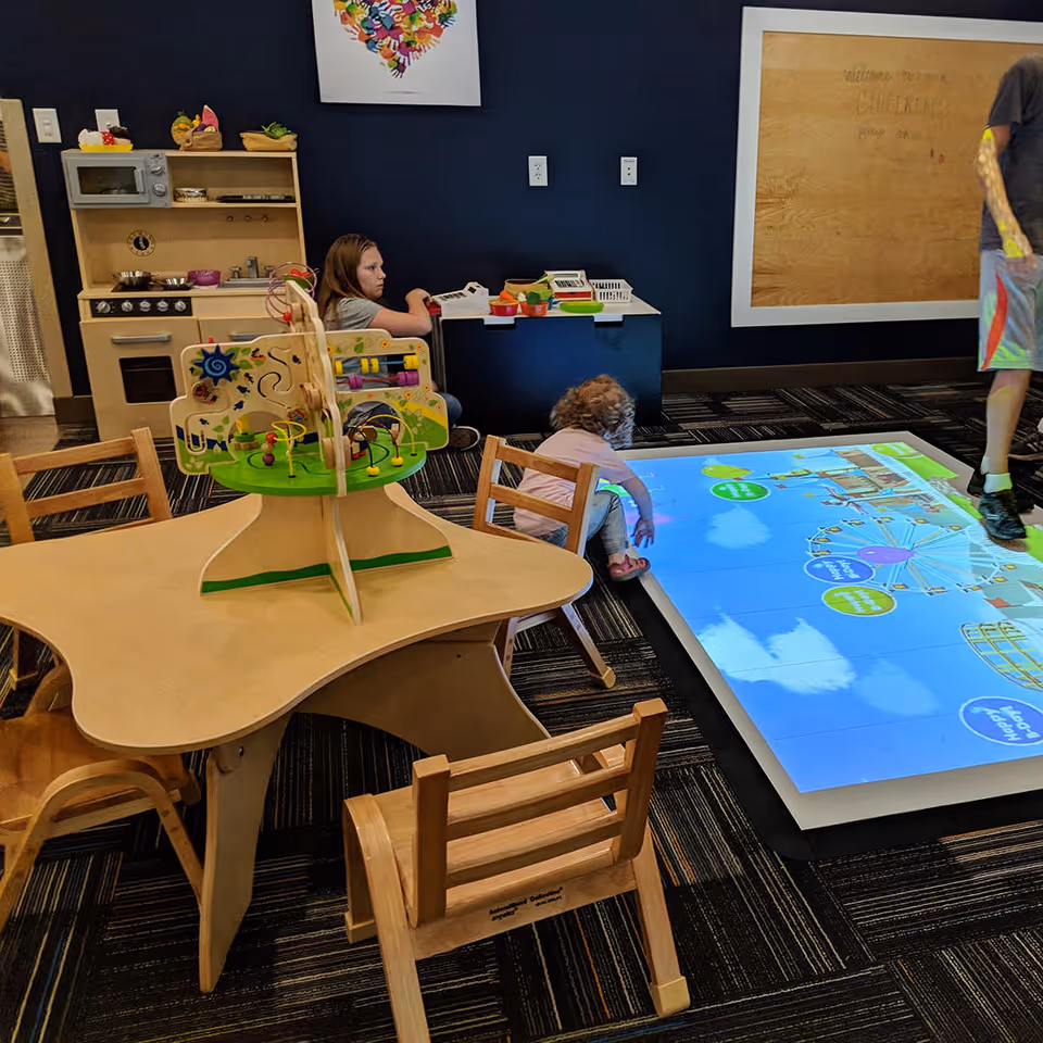 A children's play area with a wooden activity table and small chairs. A young child is interacting with a large interactive floor screen displaying colorful graphics. Another child is sitting near a small play kitchen set against a dark blue wall. An adult's legs are partially visible on the right side of the image.