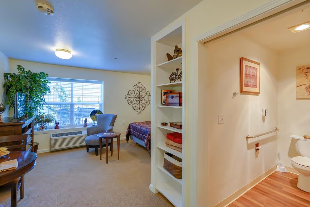 Bright studio-style senior living interior with a sitting area, shelving, bed, and an adjacent bathroom.