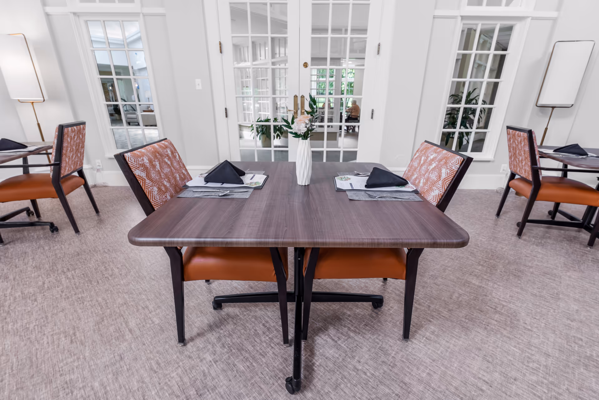 A dining area in a senior living facility with a wooden table set for two, featuring black folded napkins, placemats, and menus. The chairs have orange seats and patterned backs. In the background, there are glass-paneled double doors and windows, with plants visible through the glass.