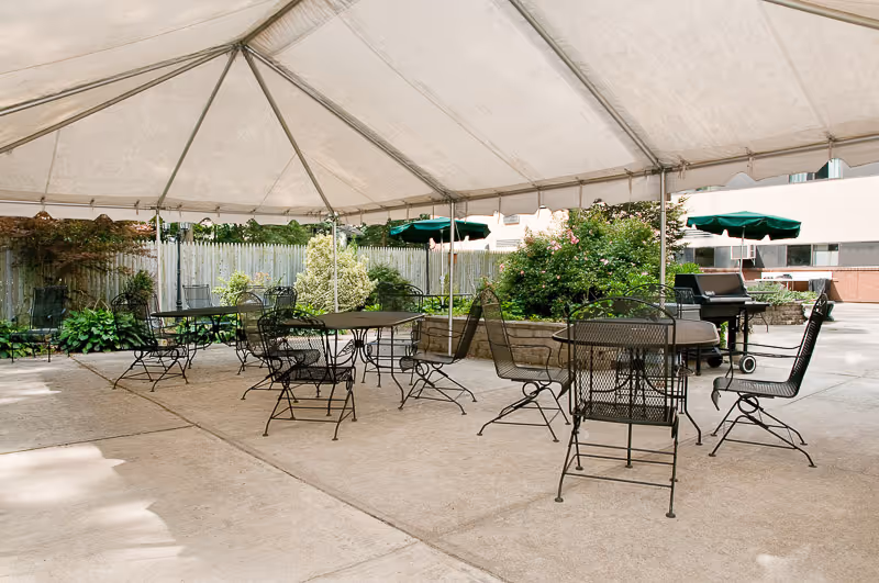 Outdoor patio area with metal tables and chairs under a large white canopy tent. There are green plants and bushes along a wooden fence in the background, and a barbecue grill is visible on the right side.