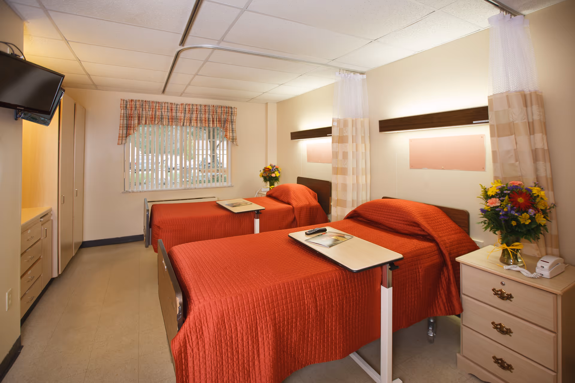 A bright and clean room with two single beds covered in red bedspreads, each with an overbed table. There is a window with vertical blinds and a plaid valance, a wall-mounted TV, a wooden dresser with a vase of colorful flowers, and a telephone. The room has light-colored walls and a tiled floor.