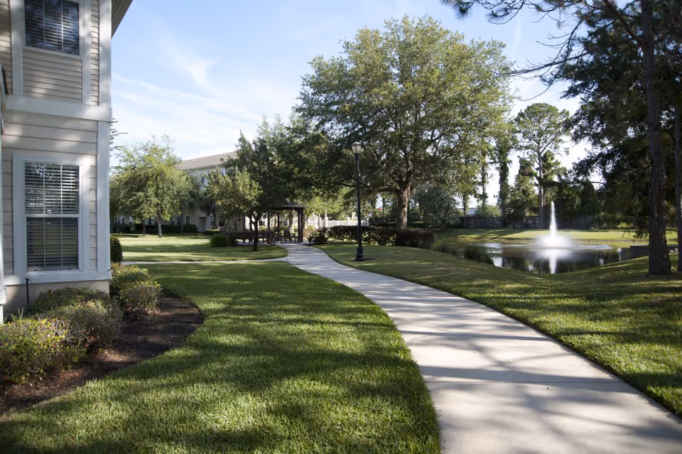 A paved walkway curves through a well-maintained grassy area with trees and shrubs. To the left is part of a building with windows, and to the right is a pond with a water fountain. A gazebo and a lamppost are visible along the path under a clear sky.