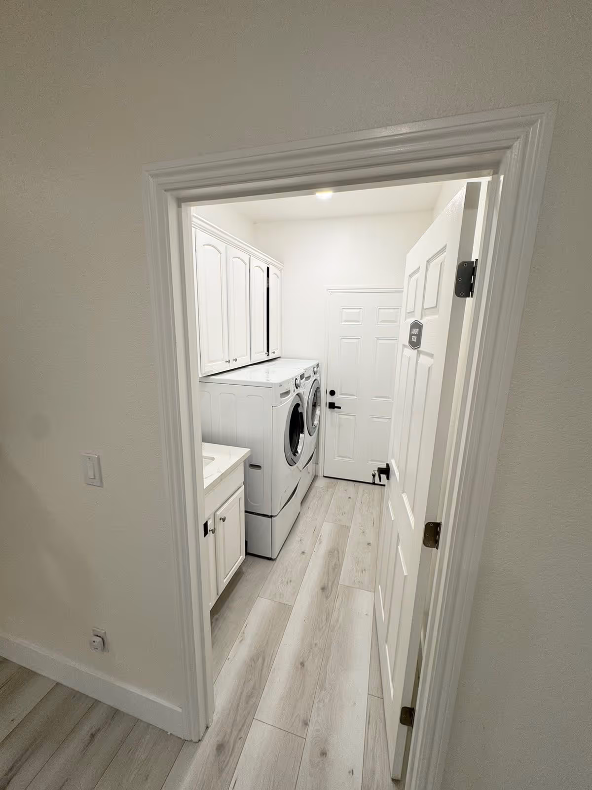 View through an open doorway into a laundry room with a white washing machine and dryer side by side, white cabinetry above and beside the machines, light wood flooring, and a white door at the end of the room.
