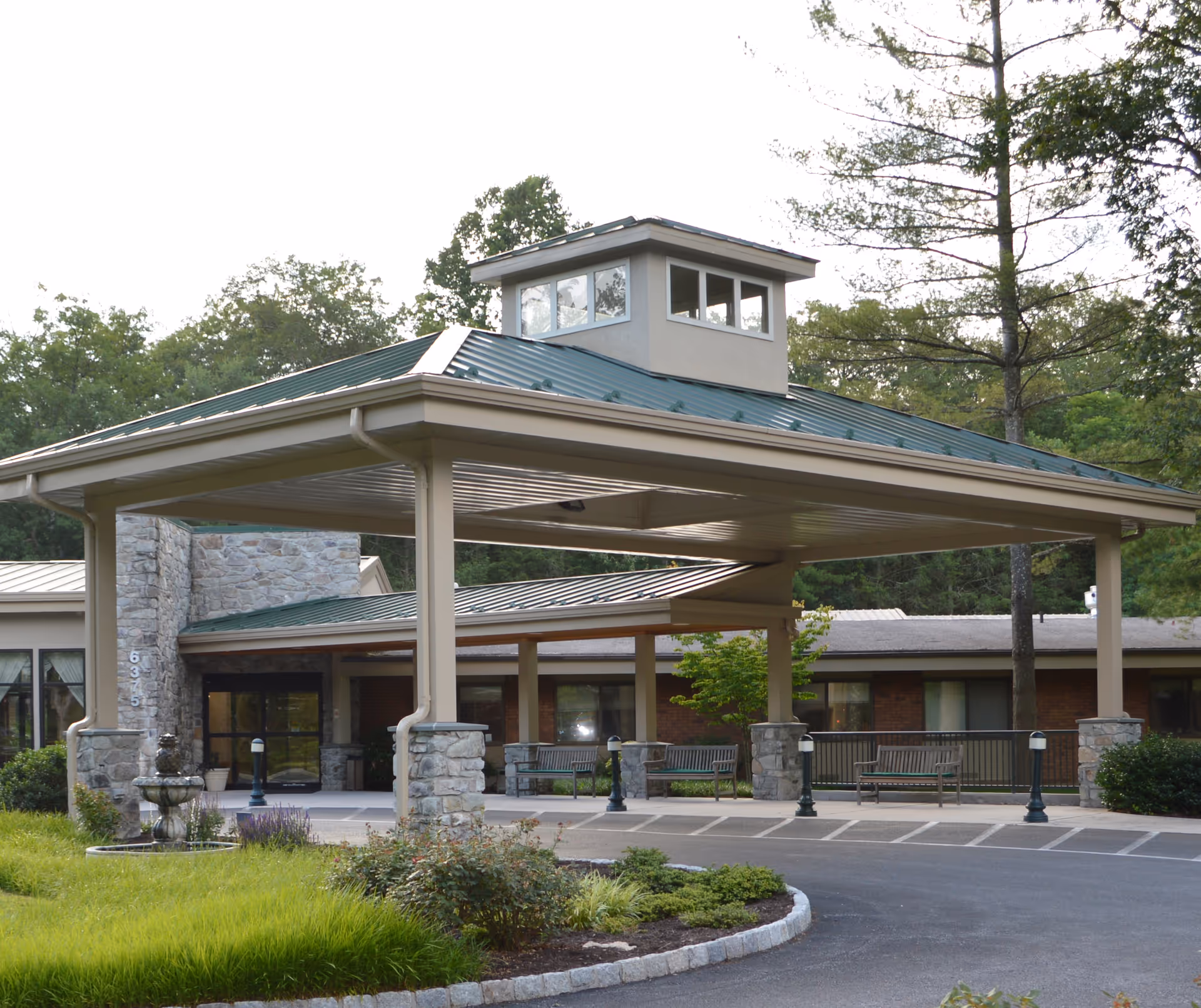 Covered porte-cochère entrance of a single-story senior living building with stone pillars, benches, and landscaping.