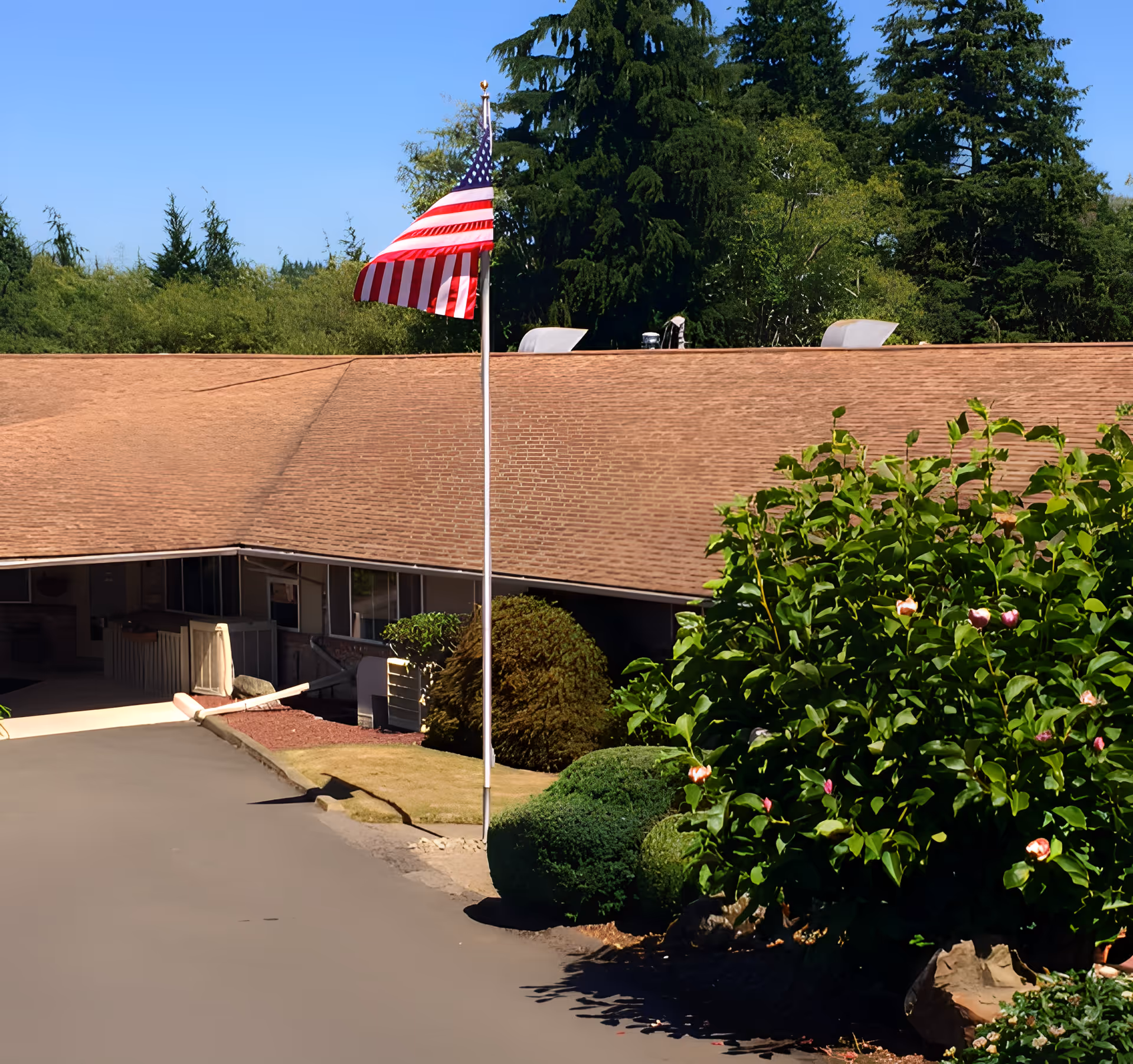 Exterior view of Grays Harbor Health & Rehabilitation Center showing a single-story building with a brown shingle roof, an American flag on a flagpole in front, and well-maintained bushes and greenery surrounding the entrance area under a clear blue sky.