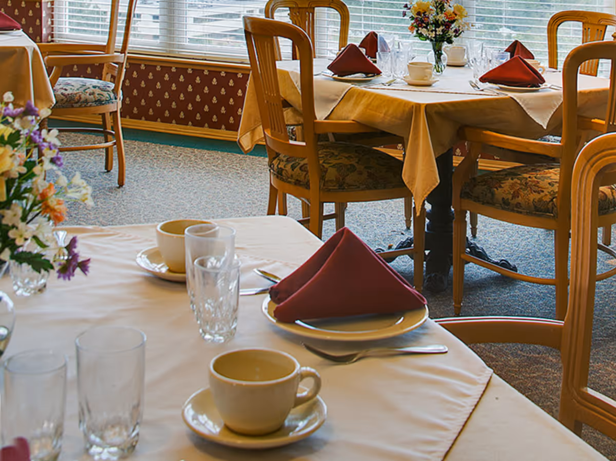 Dining room with tables set for a meal, featuring beige tablecloths, maroon folded napkins, cups, glasses, and floral centerpieces. Wooden chairs with patterned cushions surround the tables, and large windows with blinds allow natural light into the room.
