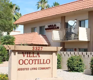 Exterior view of a two-story assisted living community building with a beige stucco finish and red tile roof. A sign in front reads '3327 Villa Ocotillo Assisted Living Community'. The building has balconies with railings and a satellite dish, surrounded by desert landscaping with bushes and palm trees.
