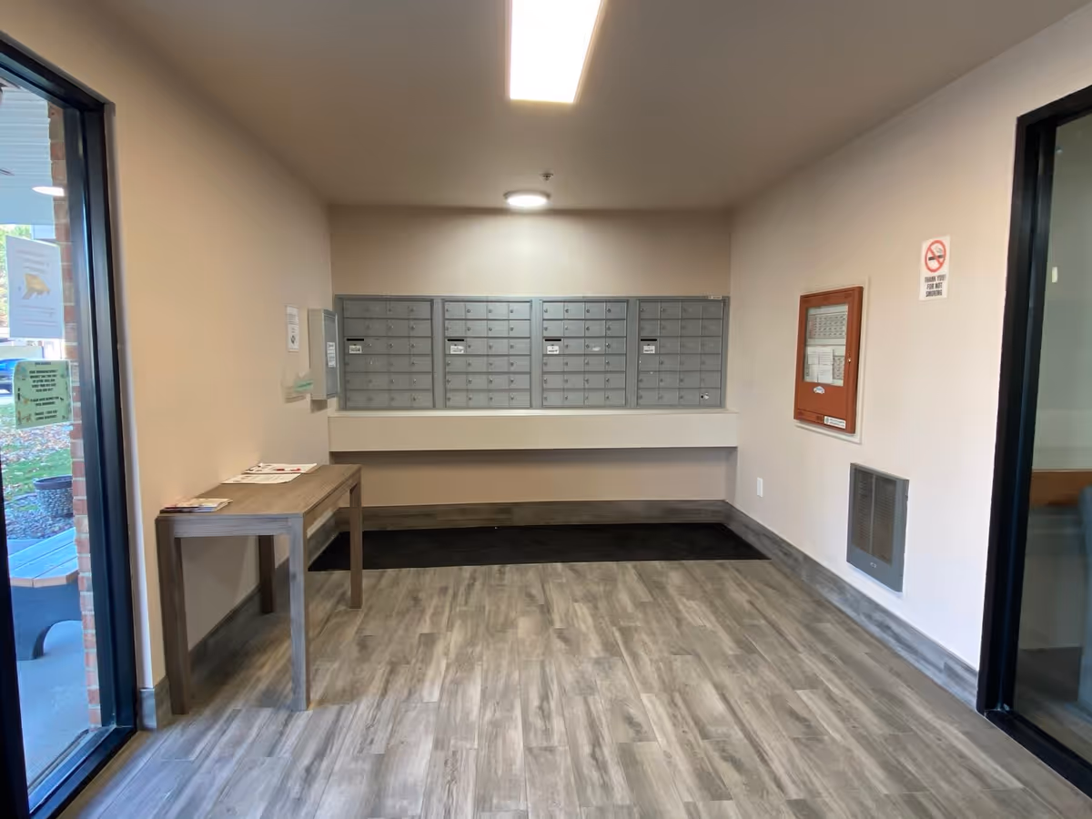 Interior view of a mailroom area in a senior living facility with multiple gray mailboxes mounted on the wall, a wooden table with some papers on it to the left, and a no smoking sign on the right wall. The floor has a wood-like finish and there are glass doors on both sides leading to other areas.