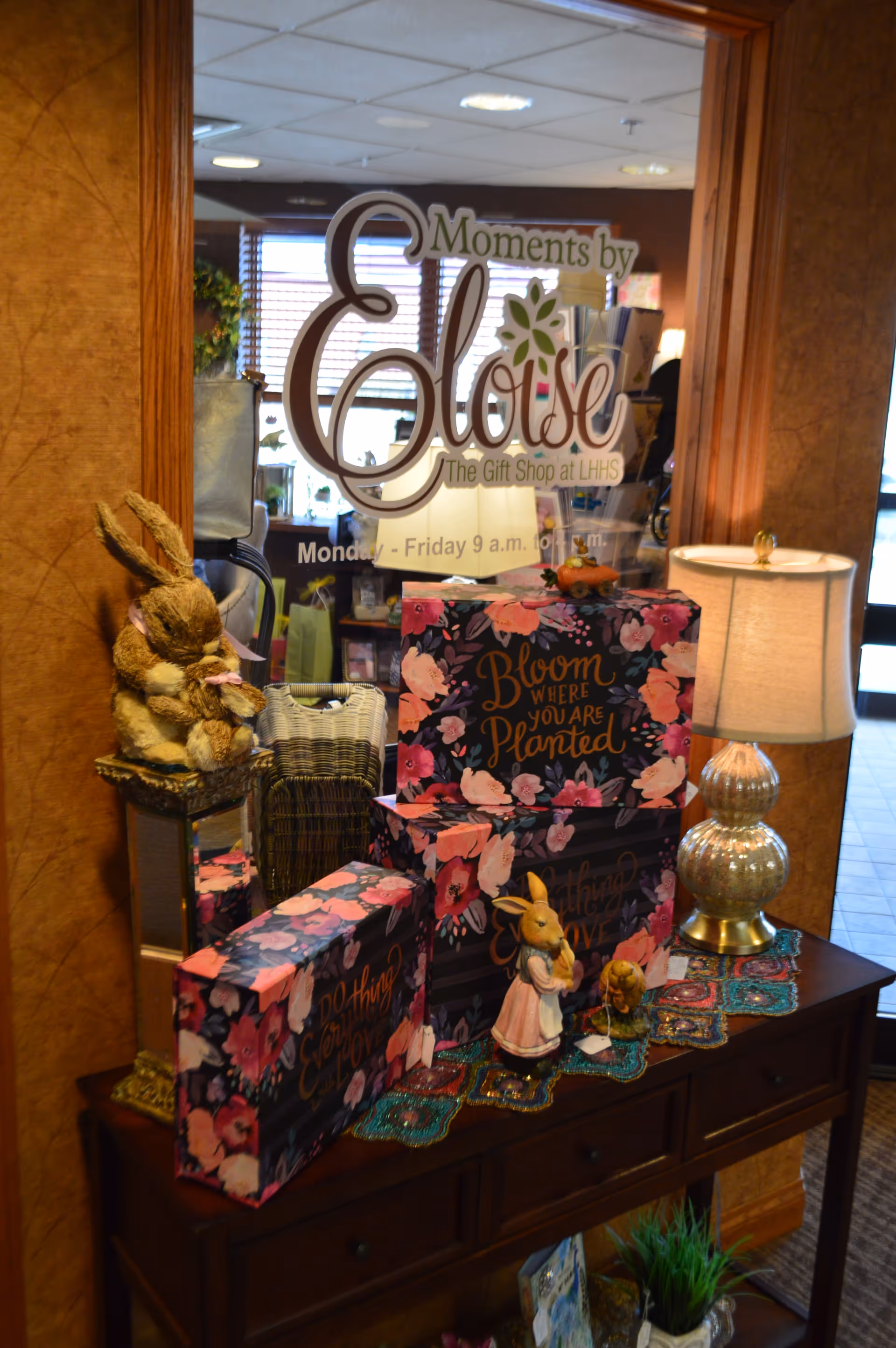 A decorative display table with floral-themed gift boxes, a lamp with a beige shade, and rabbit figurines in front of a glass door with the text 'Moments by Eloise The Gift Shop at LHHS Monday - Friday 9 a.m. to 4 p.m.'