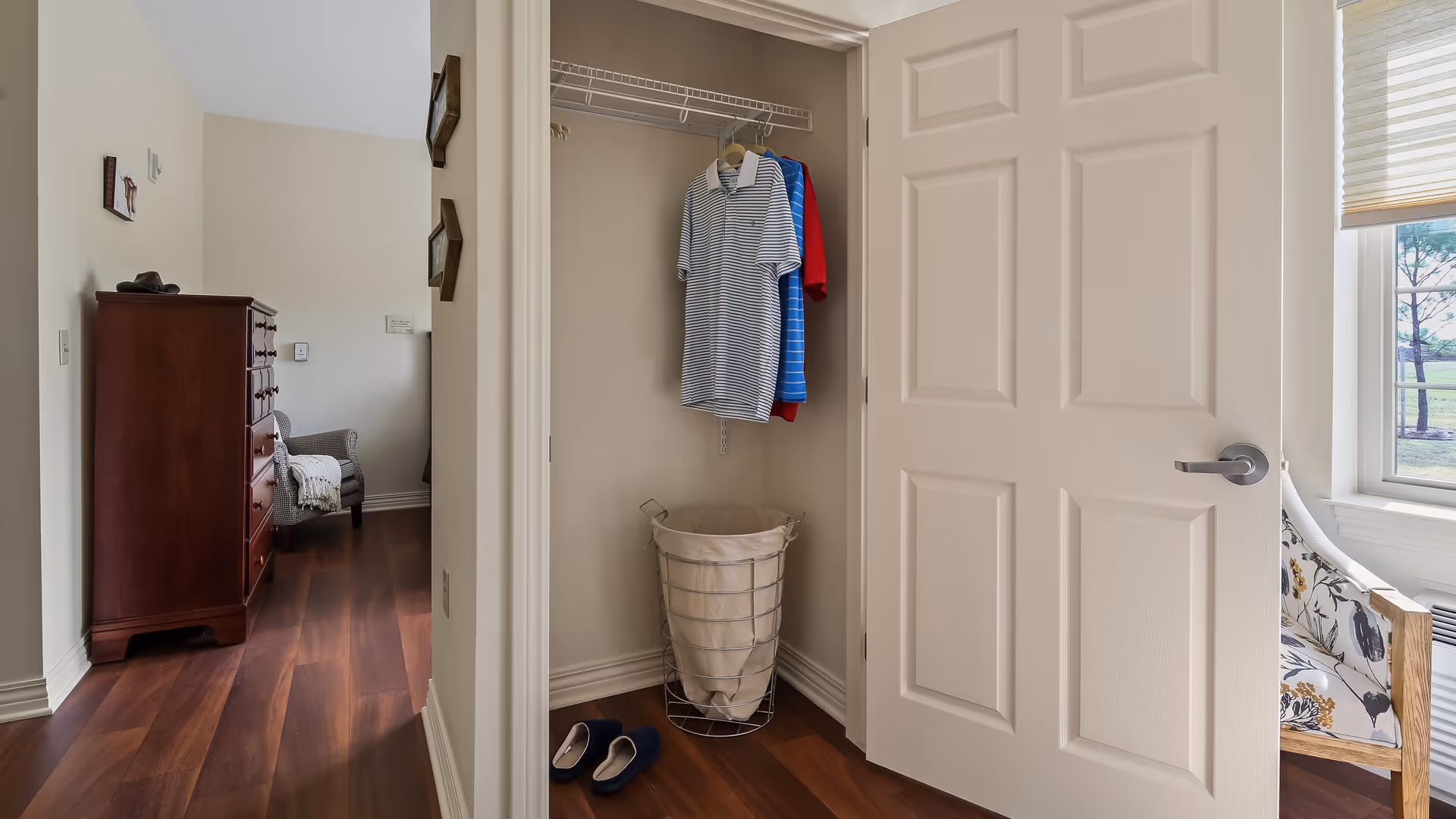View of a small closet with a few hanging shirts and a laundry basket inside. To the left, there is a hallway leading to a room with a wooden dresser and an upholstered chair. To the right, a window with a floral-patterned chair is partially visible.