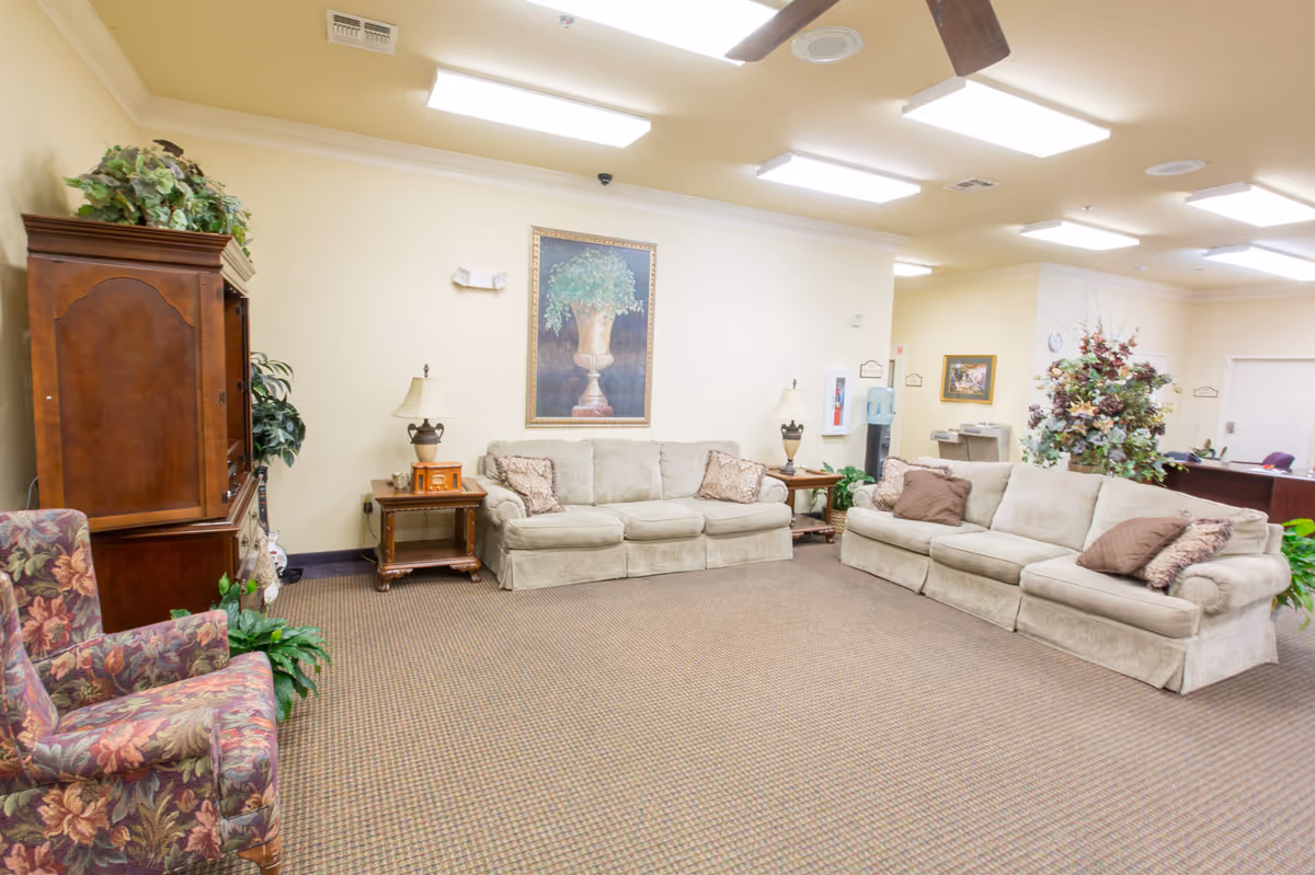 A bright communal living room with beige sofas, floral armchairs, side tables, lamps, and decorative plants and artwork.