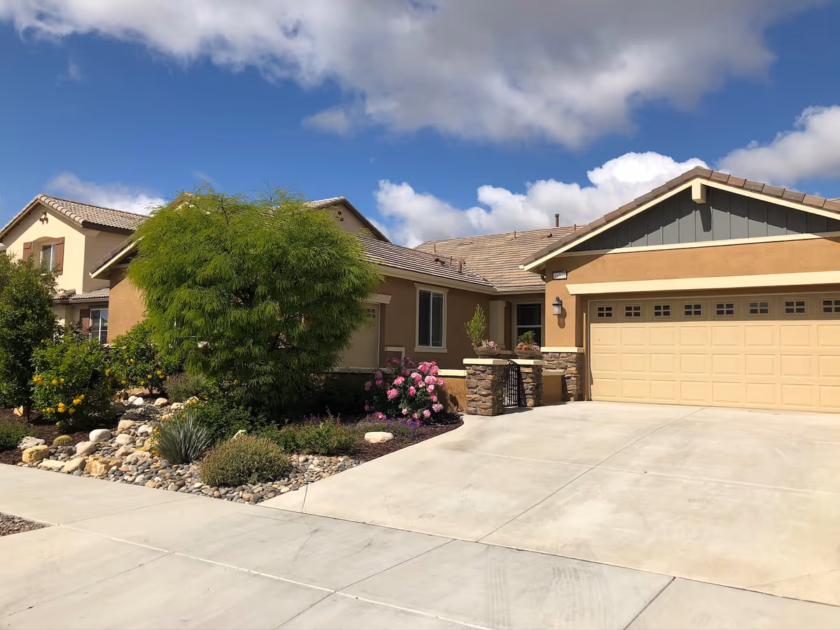 Exterior view of a single-story residential building with a beige and brown color scheme, a two-car garage, a driveway, and landscaped garden with green bushes and flowering plants under a partly cloudy blue sky.