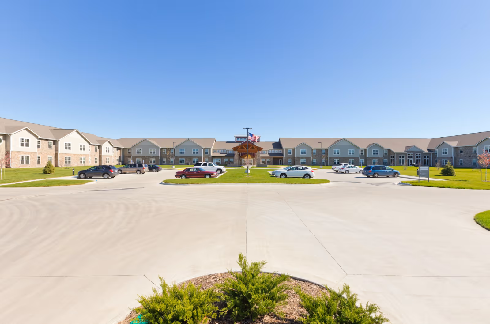 Front exterior of Prairie Vista Village showing a low-rise senior living building with a circular driveway, parked cars, and a flag at the main entrance under a clear blue sky.