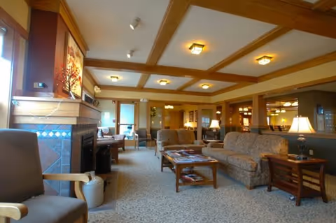 A spacious and warmly lit living room area in an assisted living community featuring patterned sofas, wooden coffee tables with magazines, a fireplace with decorative tiles, and ceiling beams with recessed lighting. The room has large windows allowing natural light and is furnished with lamps and chairs, creating a comfortable and inviting atmosphere.