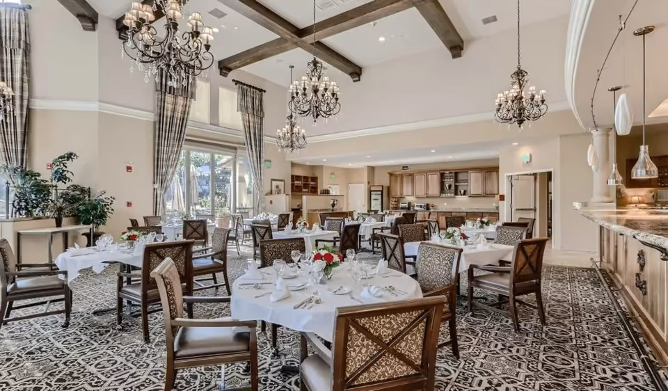 Spacious dining room with round tables set with white linens and place settings, ornate chandeliers, and a serving counter.