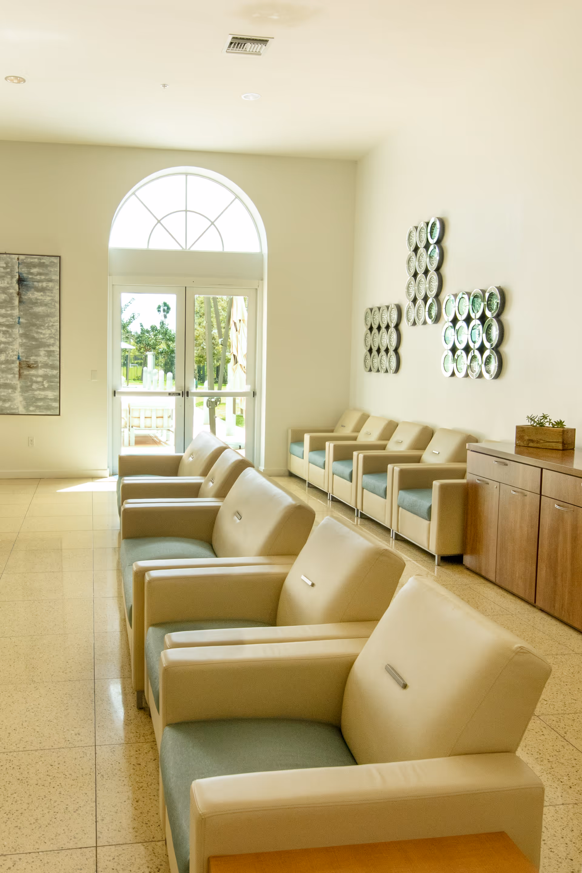 A bright waiting area with beige and light blue cushioned chairs arranged in two rows facing each other. There is a large arched window above glass double doors letting in natural light. The walls are white with decorative circular wall art and a wooden cabinet with a small plant on top.