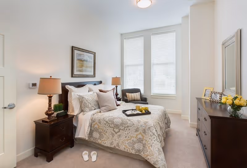 A well-lit bedroom featuring a neatly made bed with patterned bedding and multiple pillows. On either side of the bed are dark wooden nightstands with matching lamps. A pair of white slippers is placed on the carpeted floor near the bed. In the corner near two tall windows with blinds, there is a dark armchair with a striped pillow. Opposite the bed is a dark wooden dresser with a large mirror, framed photos, and a vase of yellow flowers.