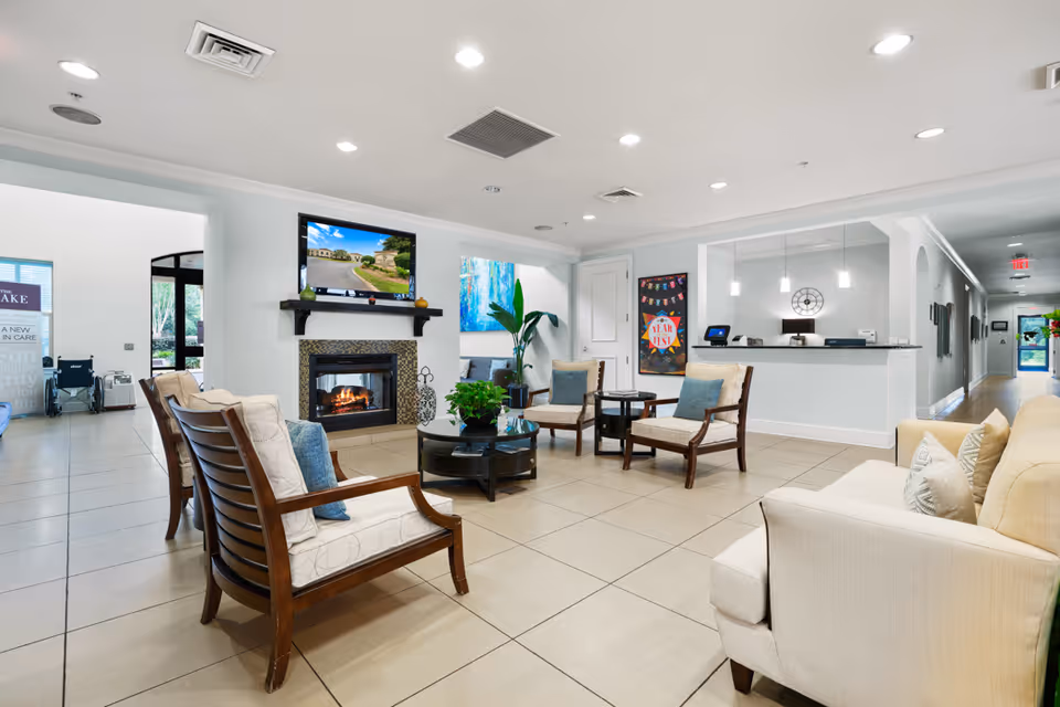 A bright and spacious senior living facility common area with beige tiled floors, several cushioned wooden chairs and a beige sofa arranged around a round black coffee table with a green plant. A fireplace with a mounted TV above it is on one wall, and a reception desk with a glass partition is visible in the background. The room is well-lit with recessed ceiling lights and has modern decor including a large plant and colorful artwork on the walls.