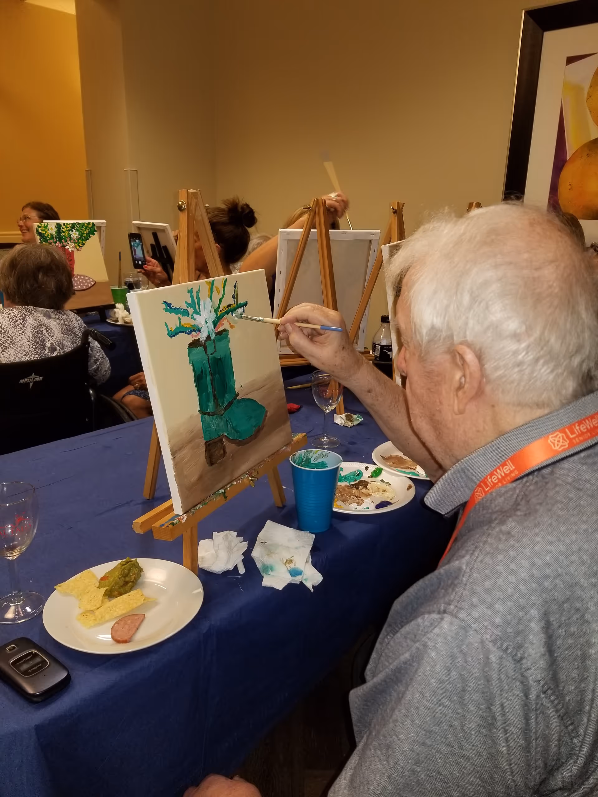An elderly man with white hair is painting a green vase with flowers on a canvas set on an easel. He is seated at a table covered with a blue tablecloth, which has a plate with chips, guacamole, and a slice of sausage, a blue plastic cup, a paint palette, and some napkins. In the background, other people are also painting on canvases set on easels.
