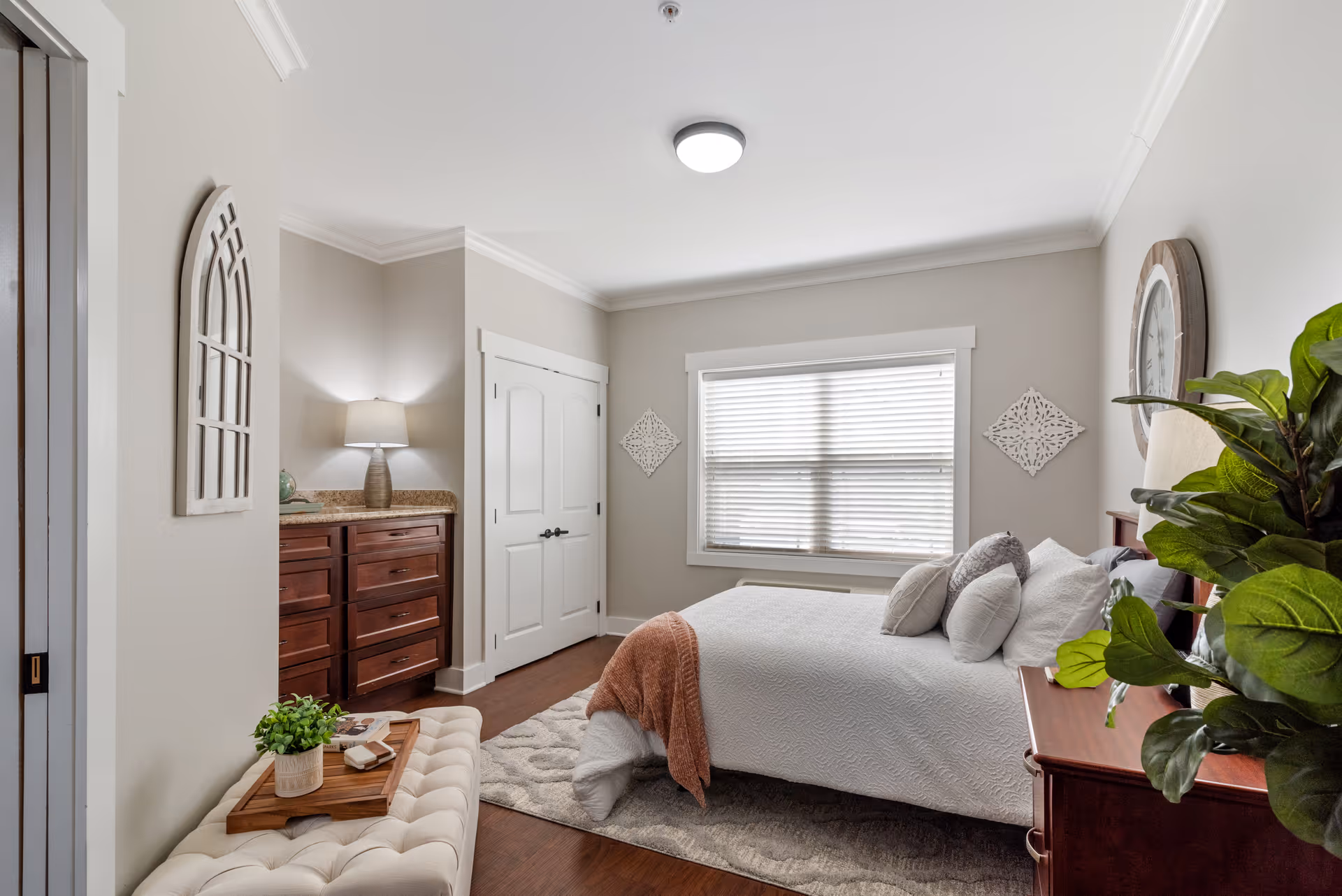 A bright and cozy bedroom with a large window covered by blinds, a bed with white bedding and multiple pillows, a wooden dresser with a lamp, a cushioned bench with a tray holding a small plant and books, decorative wall art, and a large green plant in the corner.