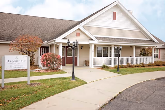 Exterior view of a single-story building with a white porch railing, two black lamp posts, and a sign that reads Brookdale Bay City. The building has a gabled roof and is surrounded by a small lawn and shrubs.