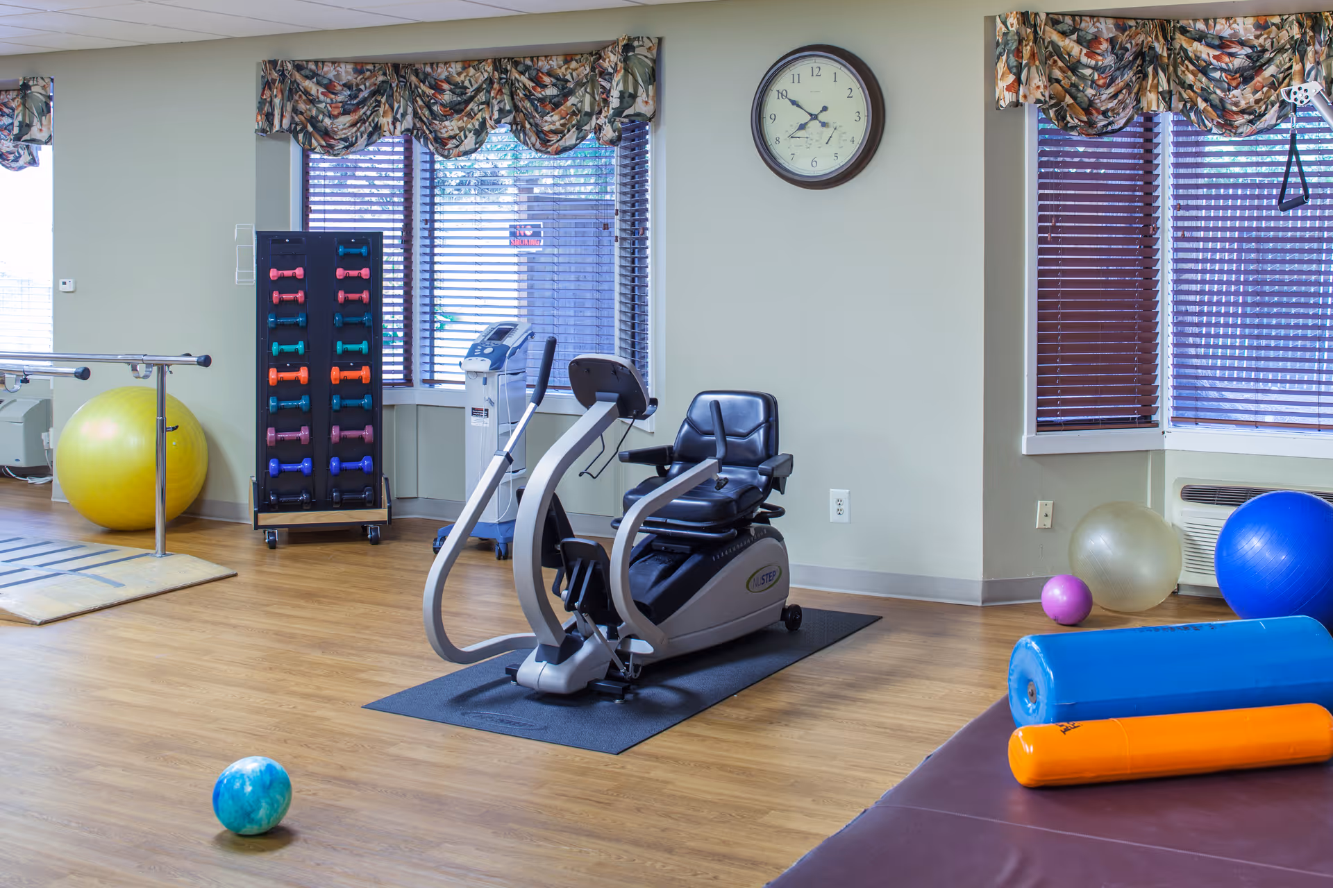 A fitness room with exercise equipment including a recumbent bike, colorful dumbbells on a rack, exercise balls, foam rollers, and parallel bars. The room has wooden flooring, large windows with floral valances, and a wall clock showing the time as 9:15.