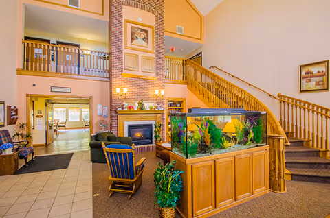 Interior view of a senior living facility lobby with a large fish tank on a wooden stand in the center. There is a staircase with wooden railings leading to an upper floor, a brick fireplace with framed artwork above it, and comfortable seating including a rocking chair and armchairs. The area is well-lit with wall sconces and natural light coming from an adjacent room.