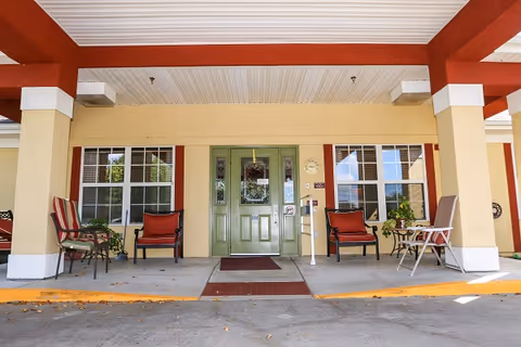 Covered entrance area of a building with a green double door in the center, flanked by two large windows with red trim. There are several chairs with cushions and small tables arranged on the concrete porch under a roof supported by beige columns with red beams.