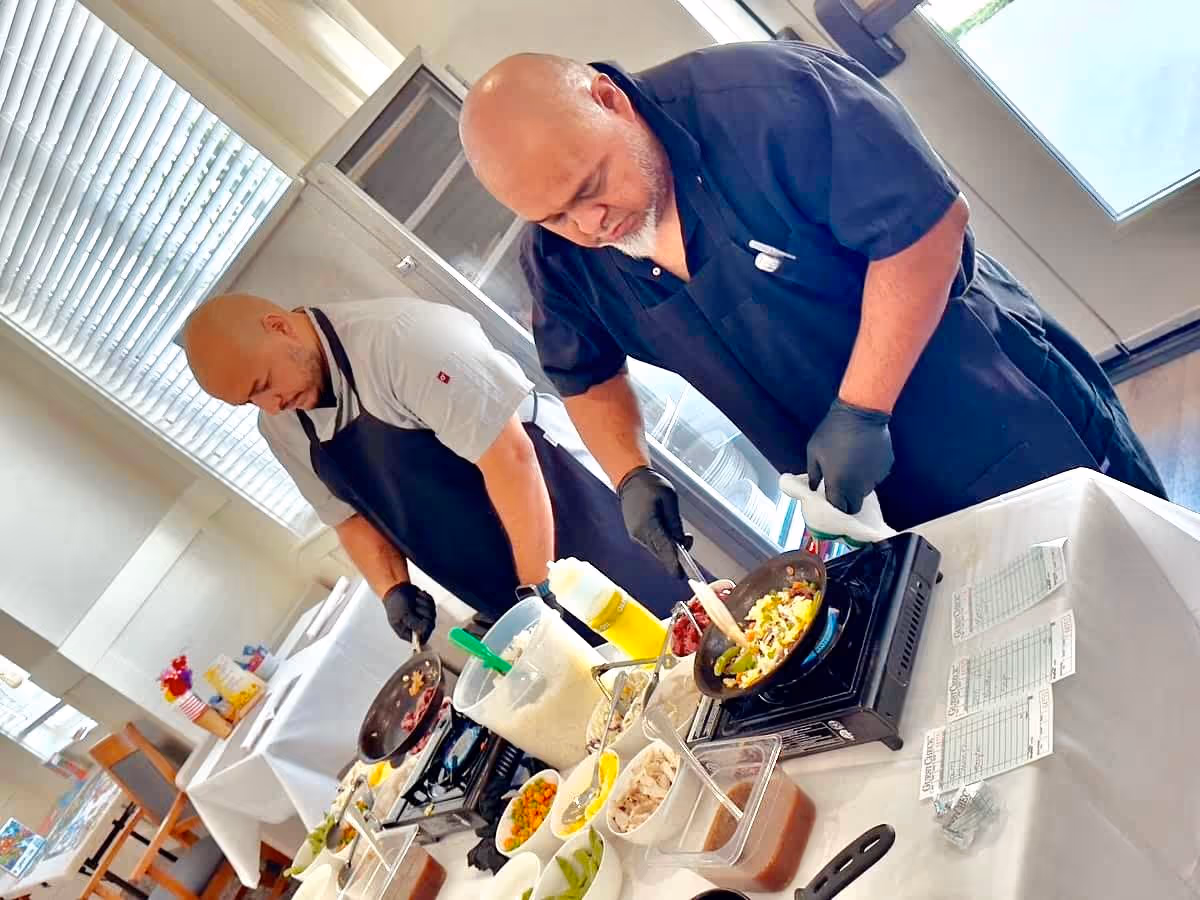 Two chefs wearing black gloves and aprons cooking food in pans on portable stoves in a bright kitchen or dining area with various ingredients and containers on the table in front of them.