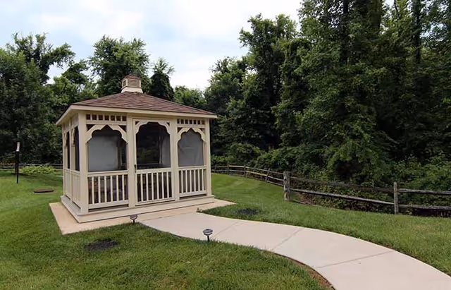 White wooden gazebo on a grassy lawn beside a curved concrete path, with trees and a split-rail fence behind it.