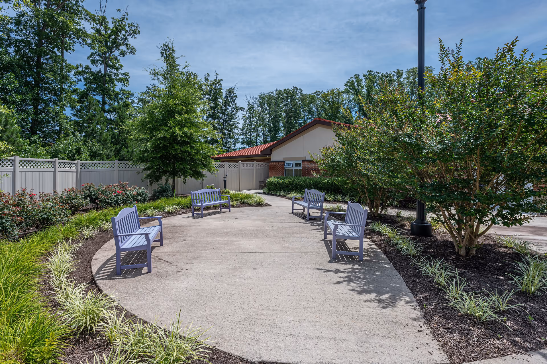 Outdoor seating area with four purple benches arranged around a circular concrete pathway, surrounded by landscaped greenery including bushes, small trees, and ornamental grasses, with a building and tall trees in the background under a partly cloudy sky.